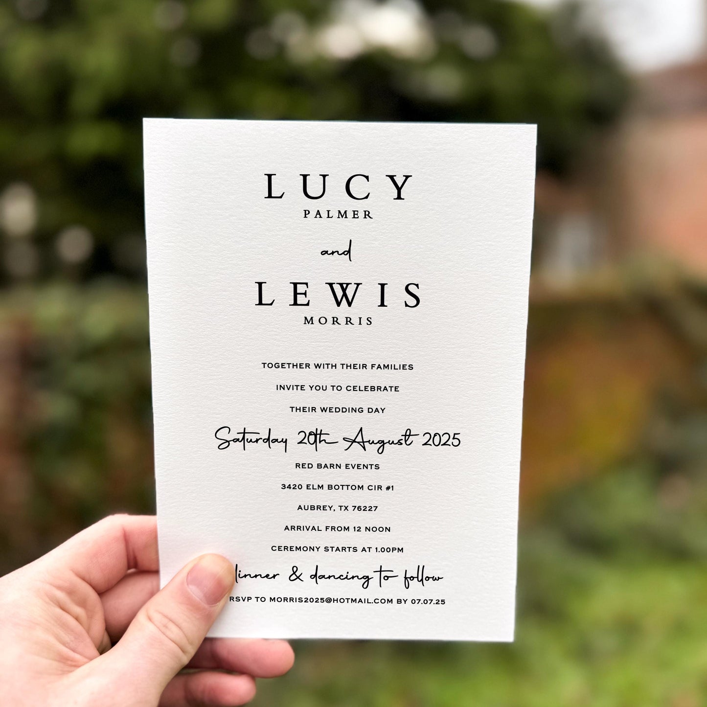 Hand holding a wedding invitation featuring bold serif names, clean black text and handwritten script accents on premium textured card.