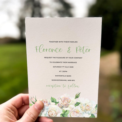 Hand holding a wedding invitation featuring soft white and blush roses with sage green foliage along the bottom edge.