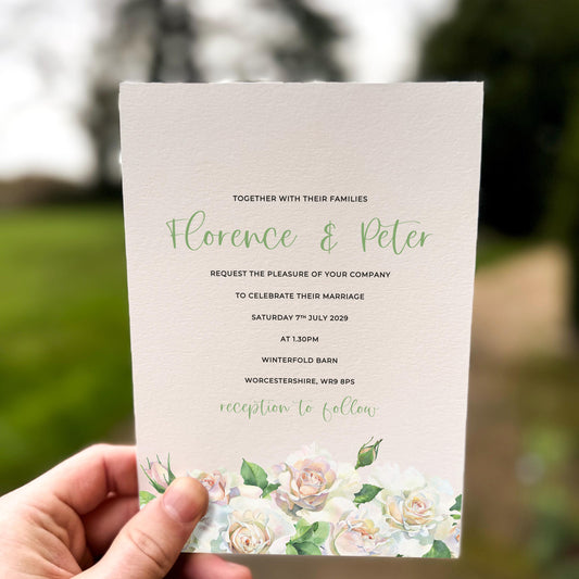 Hand holding a wedding invitation featuring soft white and blush roses with sage green foliage along the bottom edge.