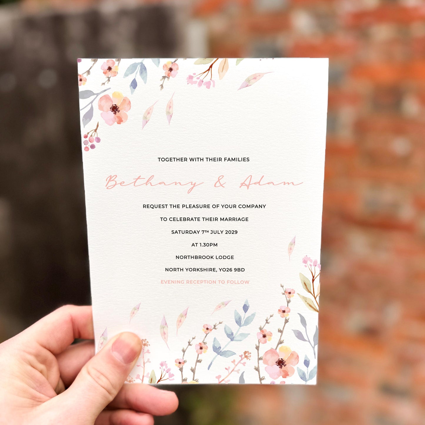 Hand holding a wedding invitation featuring delicate pastel meadow florals in blush, lavender and soft blue along the top and bottom edges.