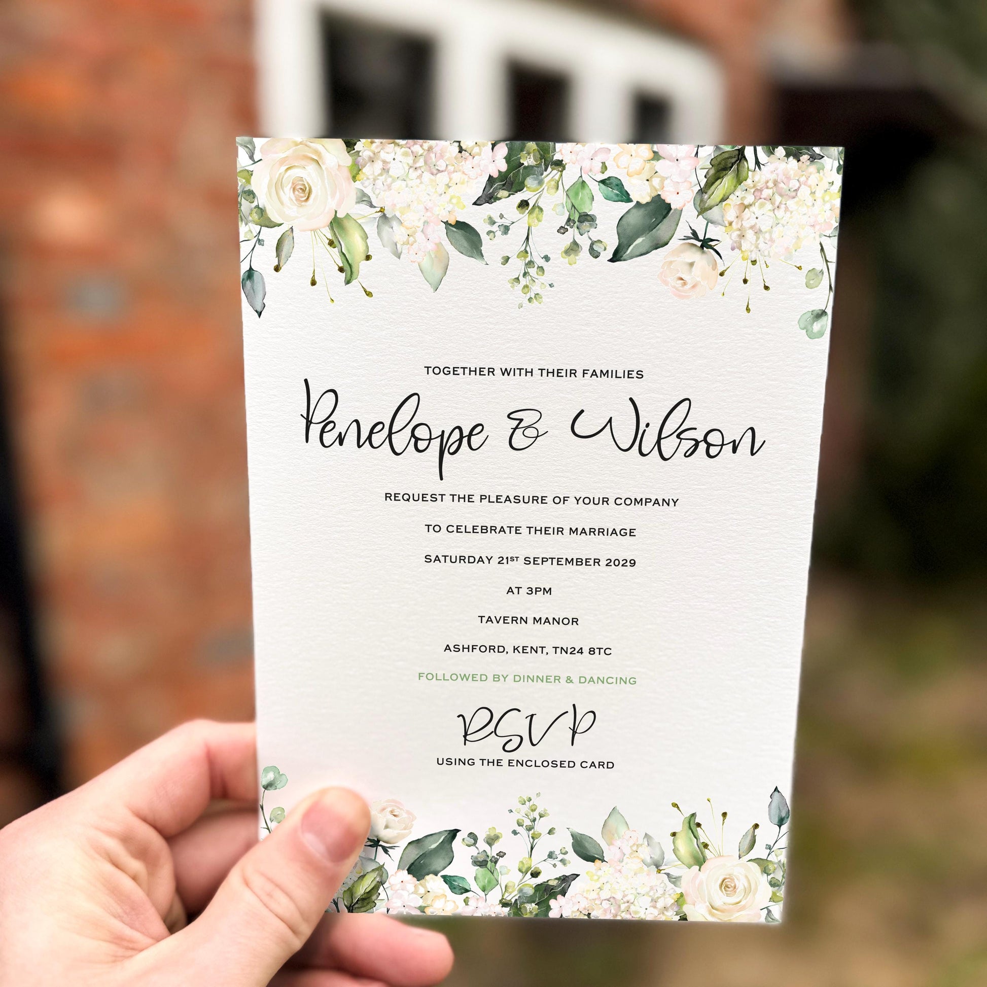 Hand holding a wedding invitation featuring white roses, hydrangeas and soft greenery arranged along the top and bottom edges.