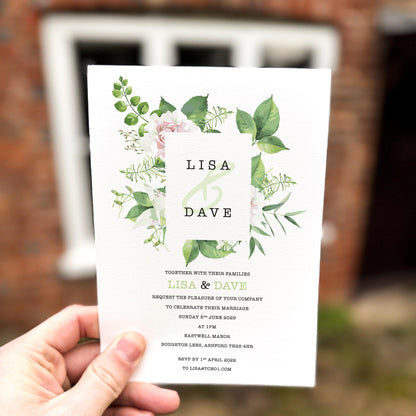 Hand holding a wedding invitation featuring a modern square frame surrounded by soft green foliage and white florals.