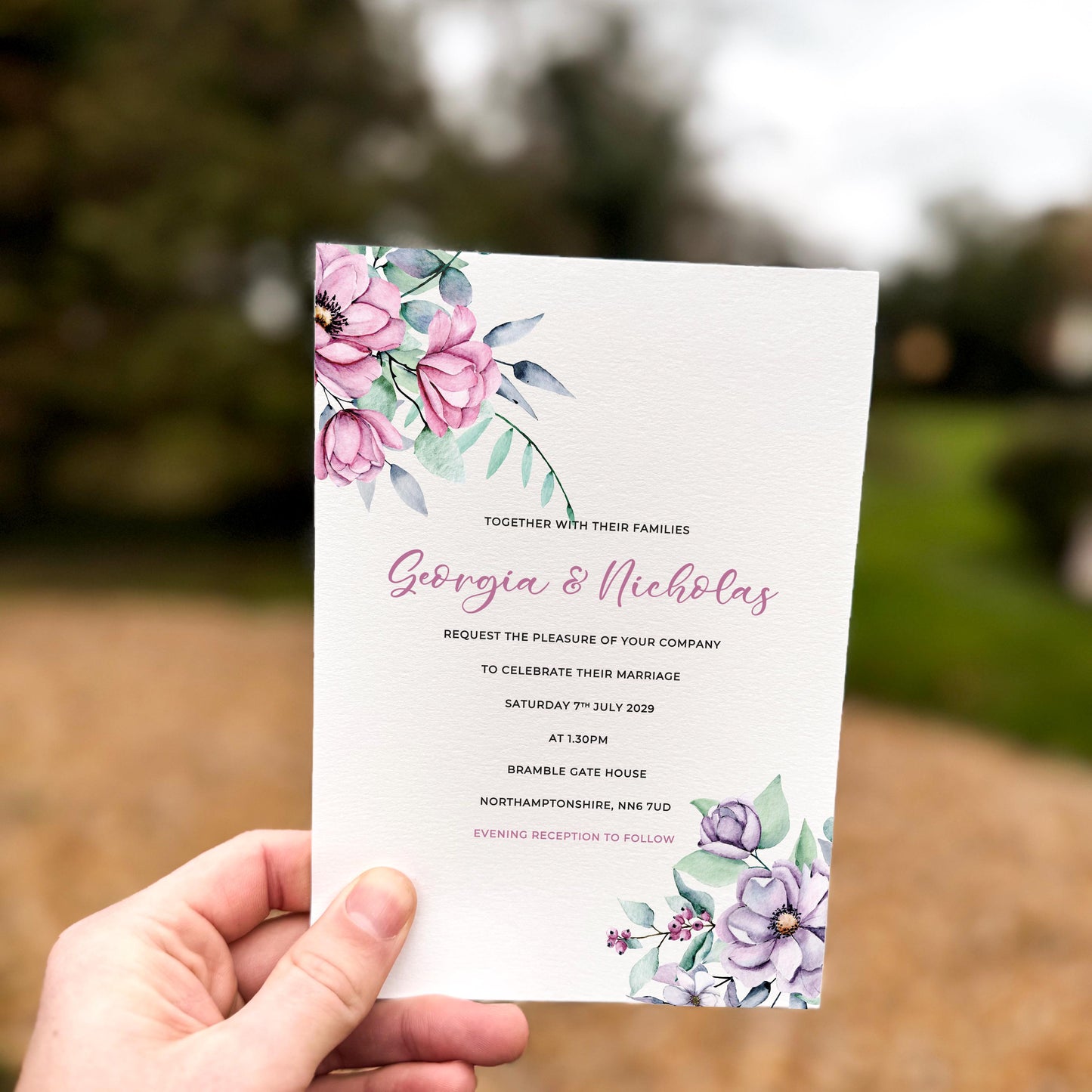 Hand holding a wedding invitation decorated with blush pink and lilac flowers, soft greenery and pastel botanical illustrations in the top left and bottom right corners.