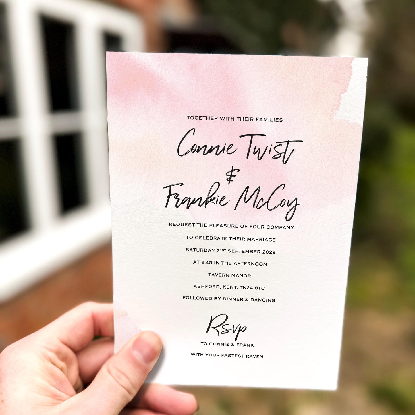 Hand holding a wedding invitation featuring a soft pink watercolour wash background with modern black script names and minimalist wedding details