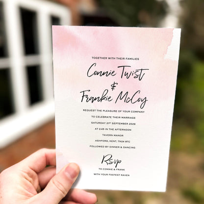 Hand holding a wedding invitation featuring a soft pink watercolour wash background with modern black script names and minimalist wedding details