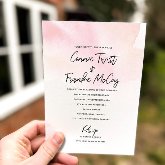 Hand holding a wedding invitation featuring a soft pink watercolour wash background with modern black script names and minimalist wedding details