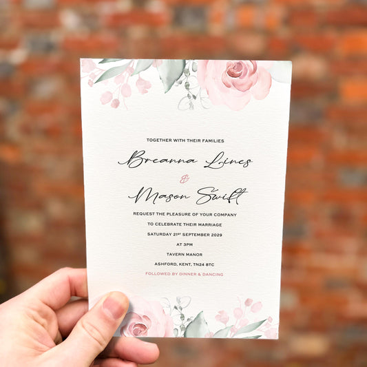Hand holding a wedding invitation with soft blush pink roses and delicate greenery at the top and bottom, printed on textured ivory card.