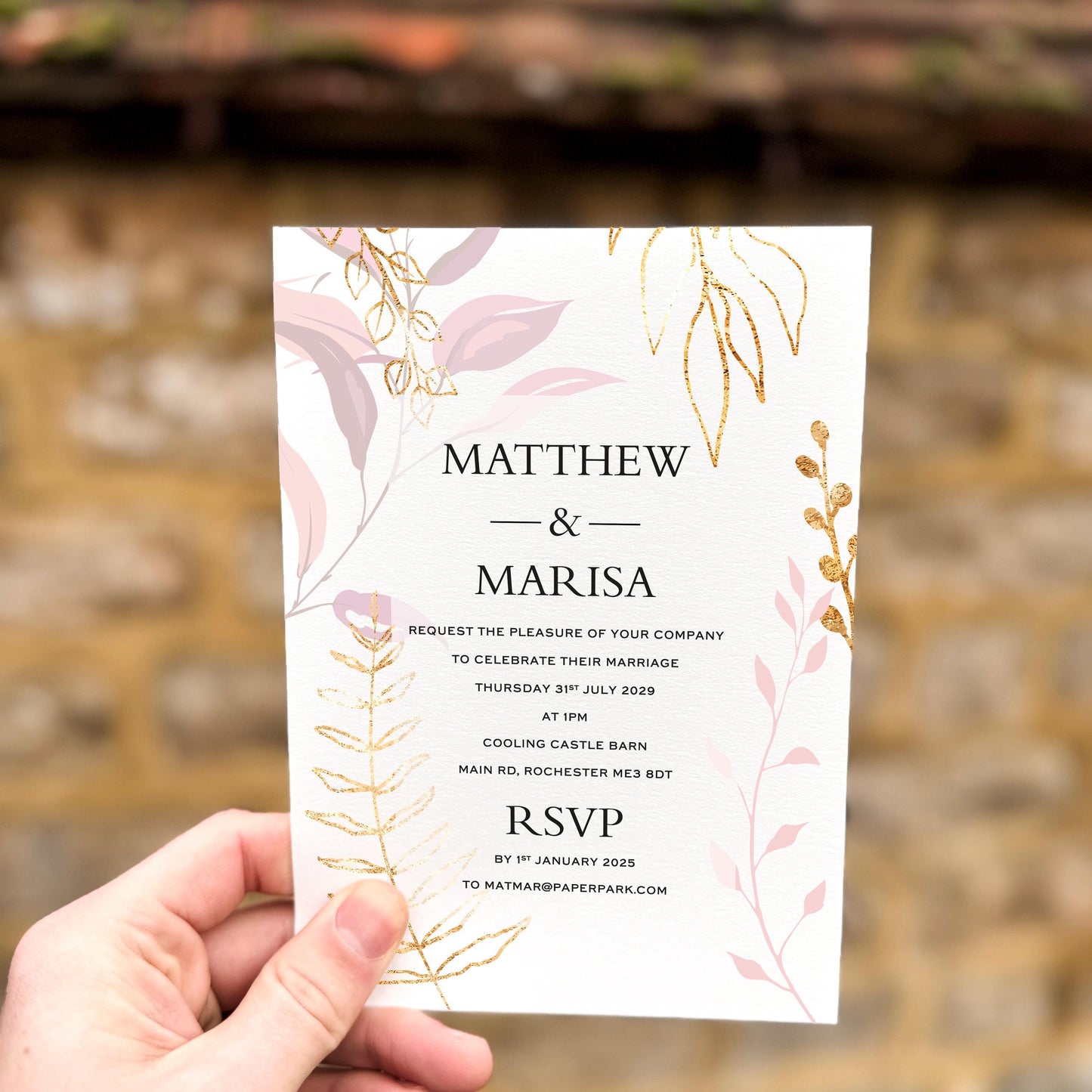 Hand holding a wedding invitation with soft pink leaves and gold botanical accents printed on textured card.