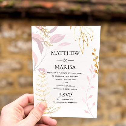 Hand holding a wedding invitation with soft pink leaves and gold botanical accents printed on textured card.