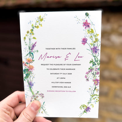 Hand holding a wedding invitation decorated with a colourful wildflower arch, featuring delicate meadow flowers framing the top and sides.