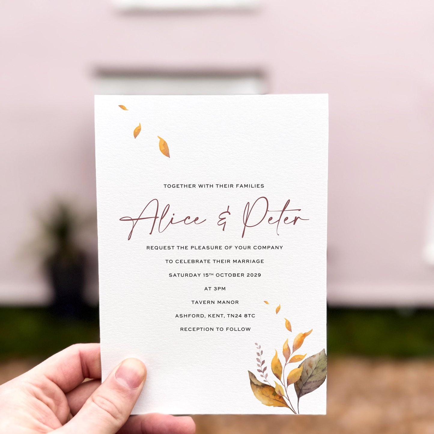 Hand holding a wedding invitation featuring delicate autumn leaves in warm golden and brown tones, printed on textured card with minimal botanical accents.