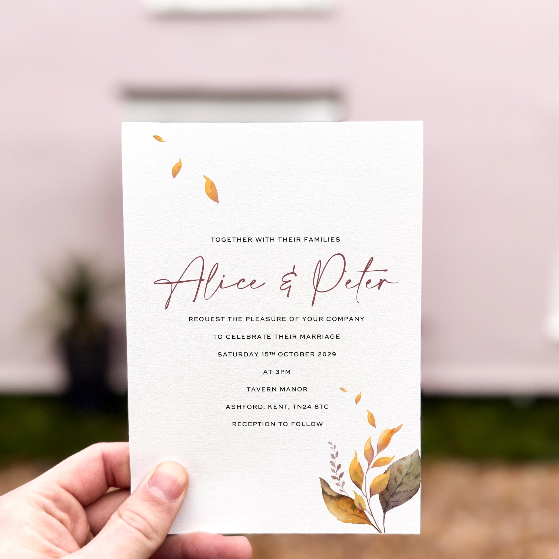 Hand holding a wedding invitation featuring delicate autumn leaves in warm golden and brown tones, printed on textured card with minimal botanical accents.