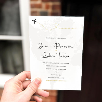 Hand holding a wedding invitation featuring a soft grey world map background with a dotted gold airplane path forming a heart.