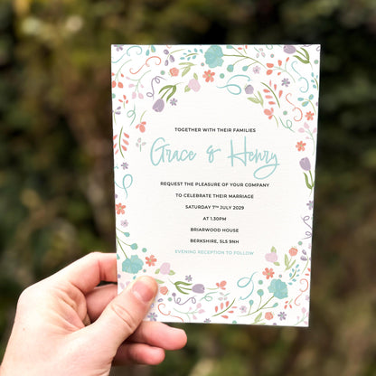 Hand holding a wedding invitation featuring a colourful pastel floral border with whimsical garden flowers and delicate illustrated leaves.
