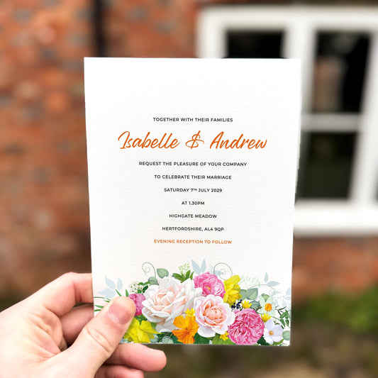 Hand holding a spring wedding invitation with bright yellow, pink and white flowers arranged along the bottom edge.