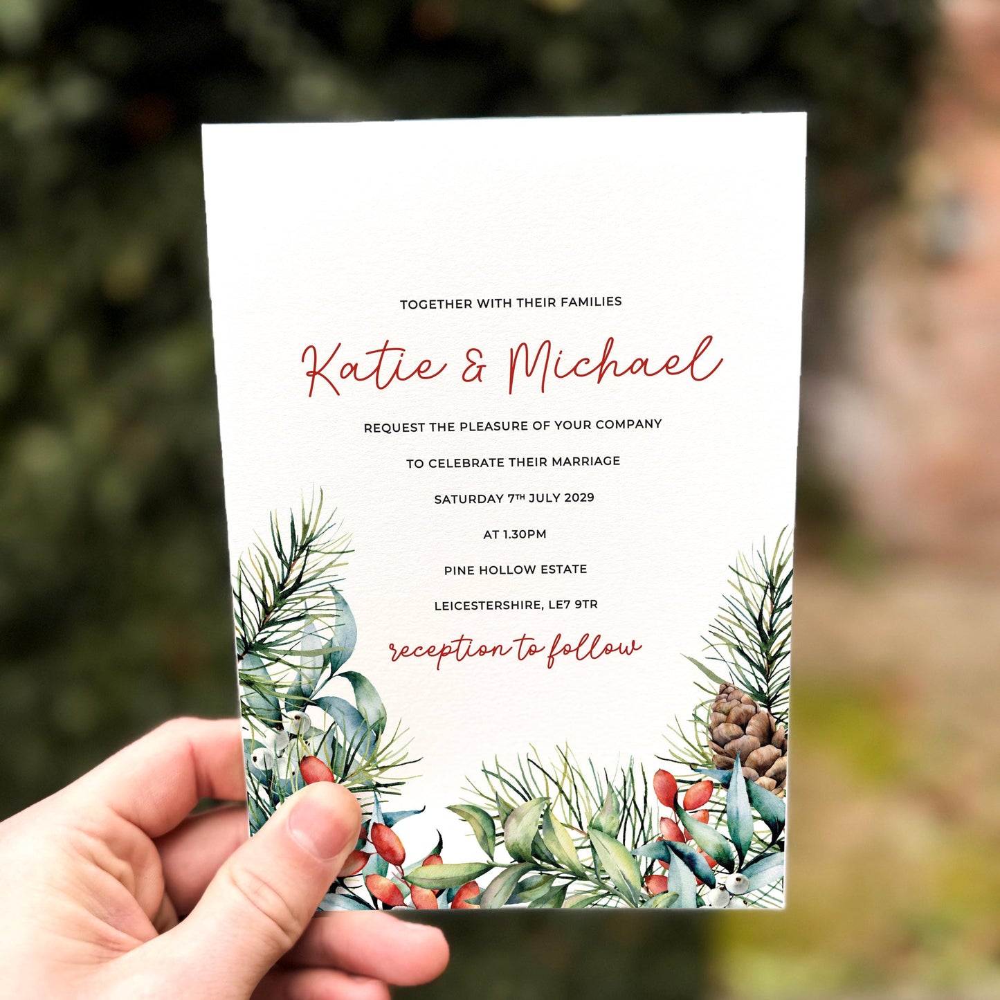 Hand holding a winter wedding invitation featuring pine branches, red berries, eucalyptus leaves and pinecones arranged along the bottom edge on textured white card.