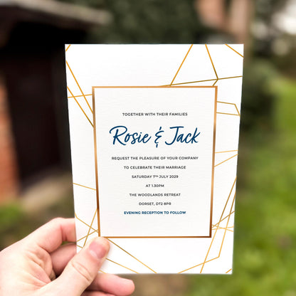 Hand holding a wedding invitation featuring modern gold geometric linework framing elegant navy text.