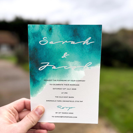 Hand holding a wedding invitation featuring a bold teal watercolour wash fading into white with modern script text.