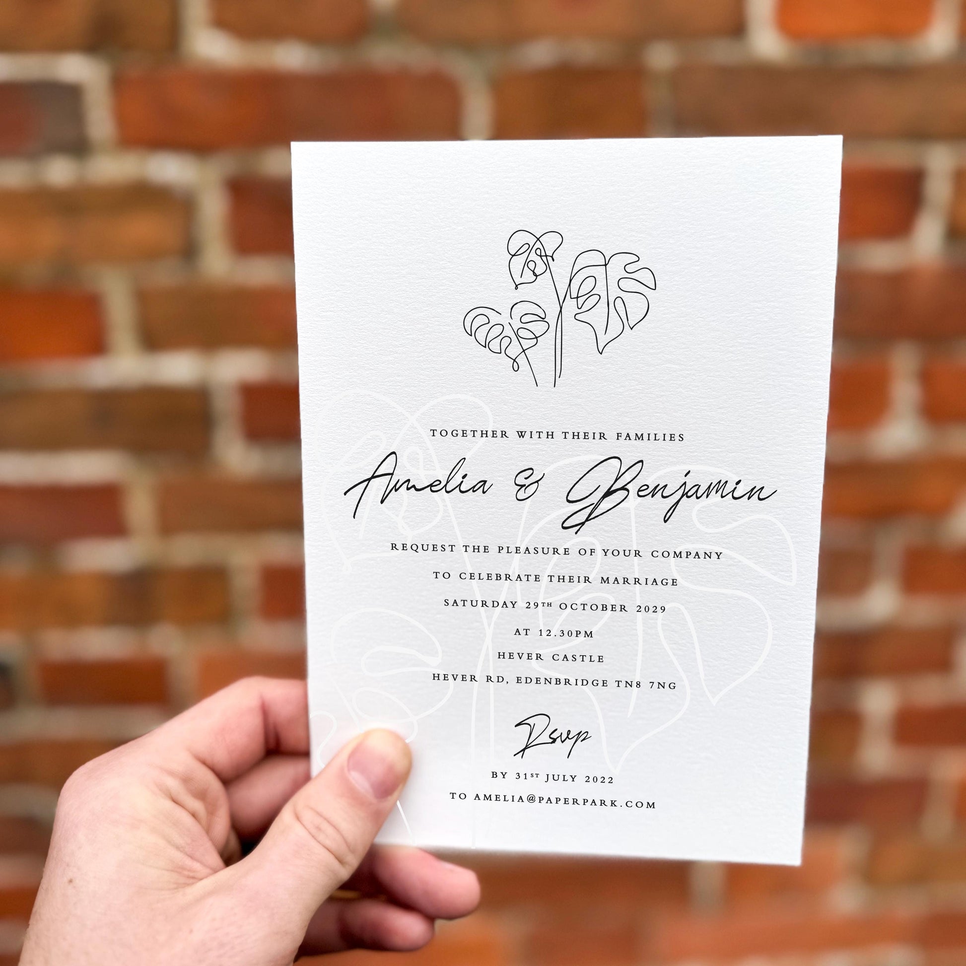 Hand holding a wedding invitation featuring minimalist monstera leaf line art with elegant black script text.