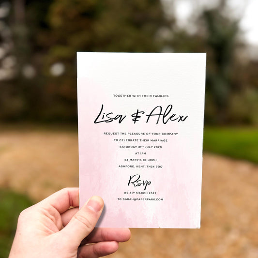 Hand holding a wedding invitation featuring a soft blush pink watercolour wash with modern black script text.