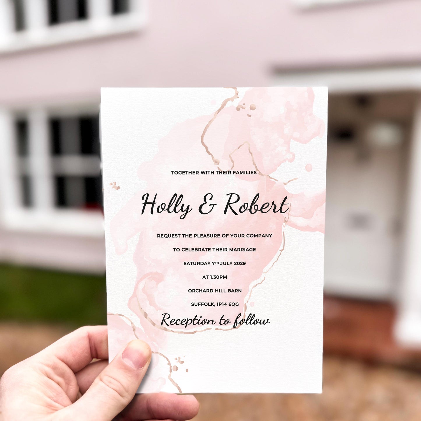 Hand holding a wedding invitation featuring soft blush pink abstract ink-wash shapes with elegant black script text.