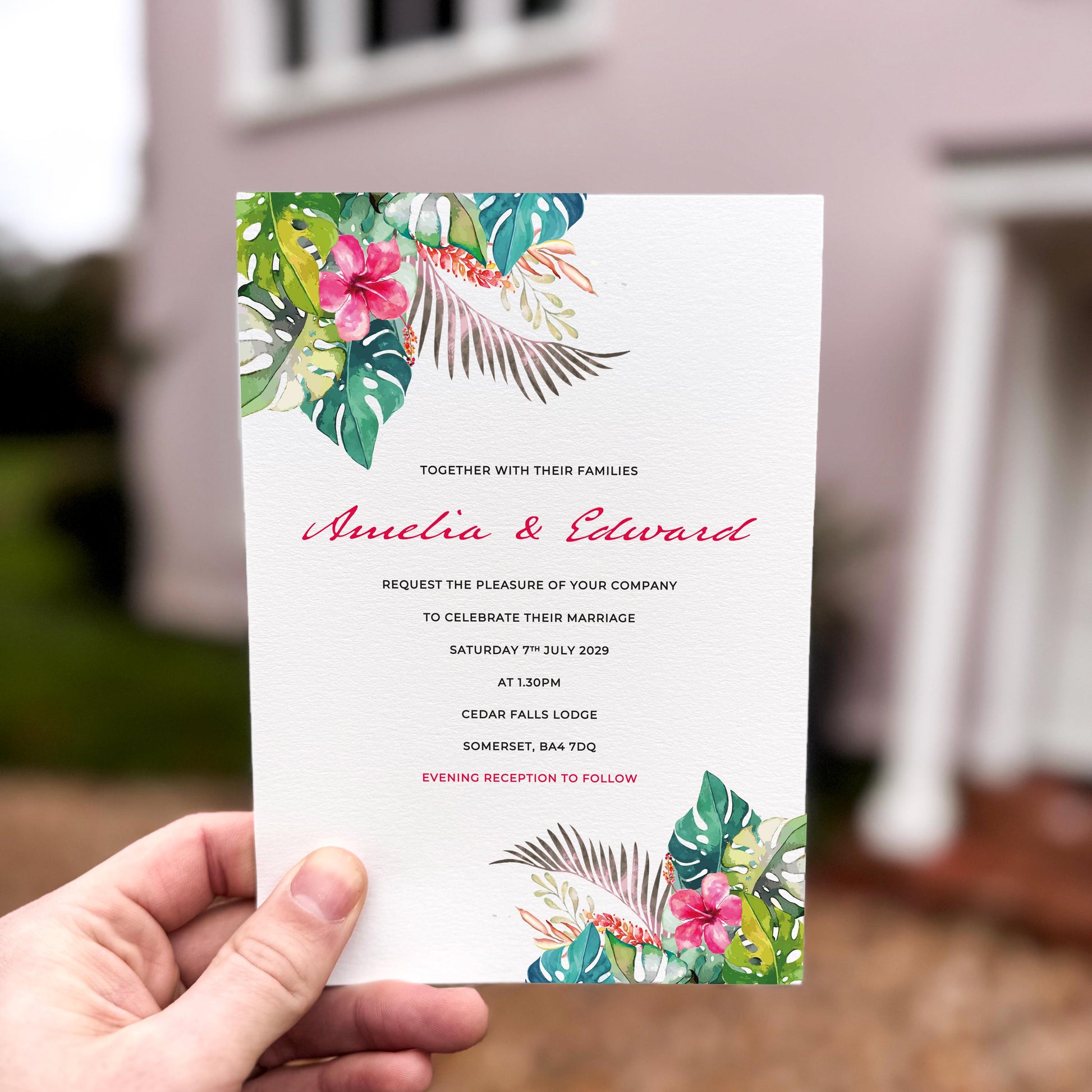 Hand holding a wedding invitation featuring vibrant tropical leaves and pink florals at the top and bottom edges.