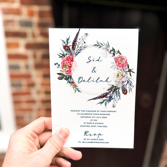 Hand holding a wedding invitation featuring a circular wreath of boho florals, feathers and foliage with deep pink and navy accents.