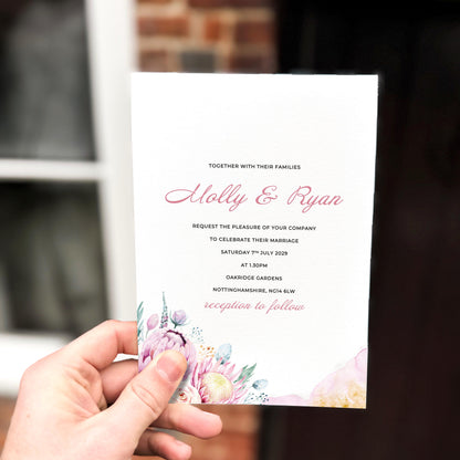 Hand holding a wedding invitation featuring a soft pastel floral cluster in blush, lilac and peach tones at the bottom edge.