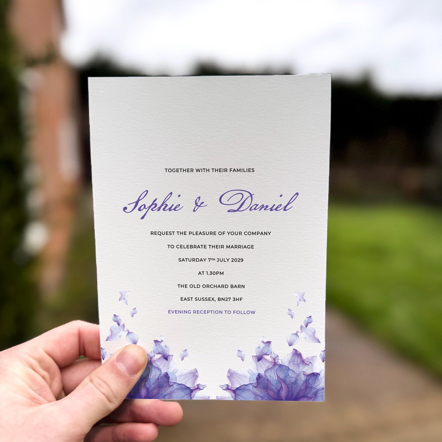Hand holding a wedding invitation featuring soft purple watercolour petals rising from the bottom edge with elegant matching script text.