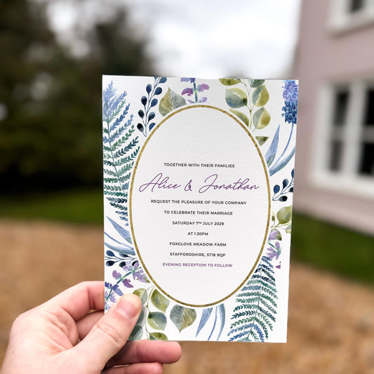 Hand holding a wedding invitation featuring an oval gold frame surrounded by wild greenery, ferns and blue–purple foliage.
