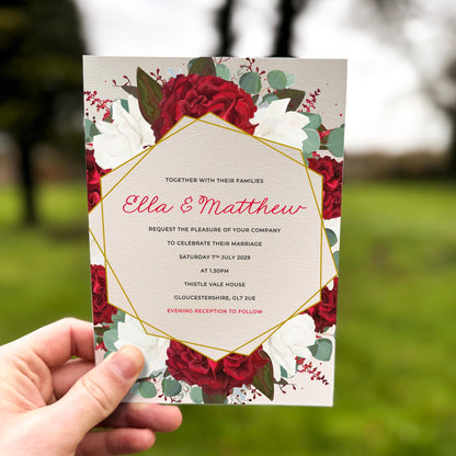 Hand holding a wedding invitation featuring deep red and white roses surrounding a modern gold geometric frame.