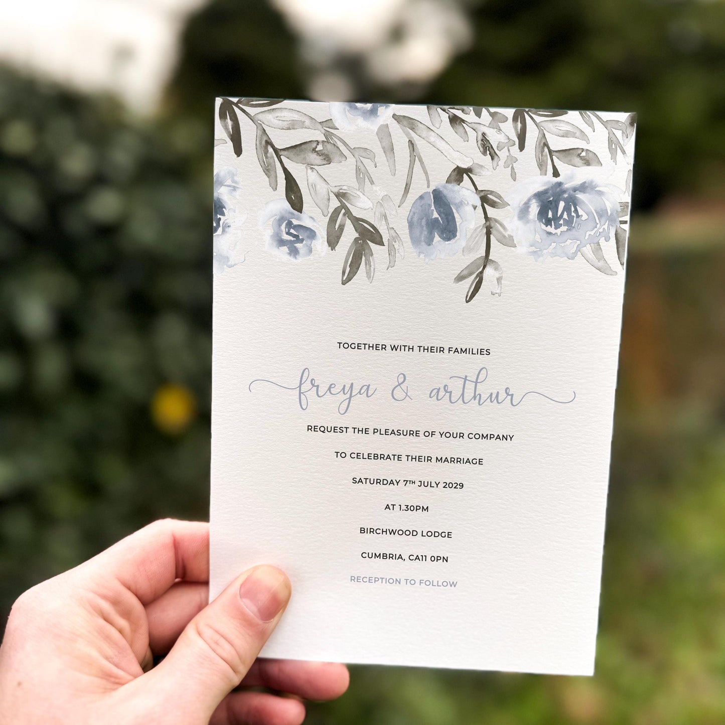 Hand holding a wedding invitation featuring soft blue-grey watercolour florals and leafy foliage across the top border.