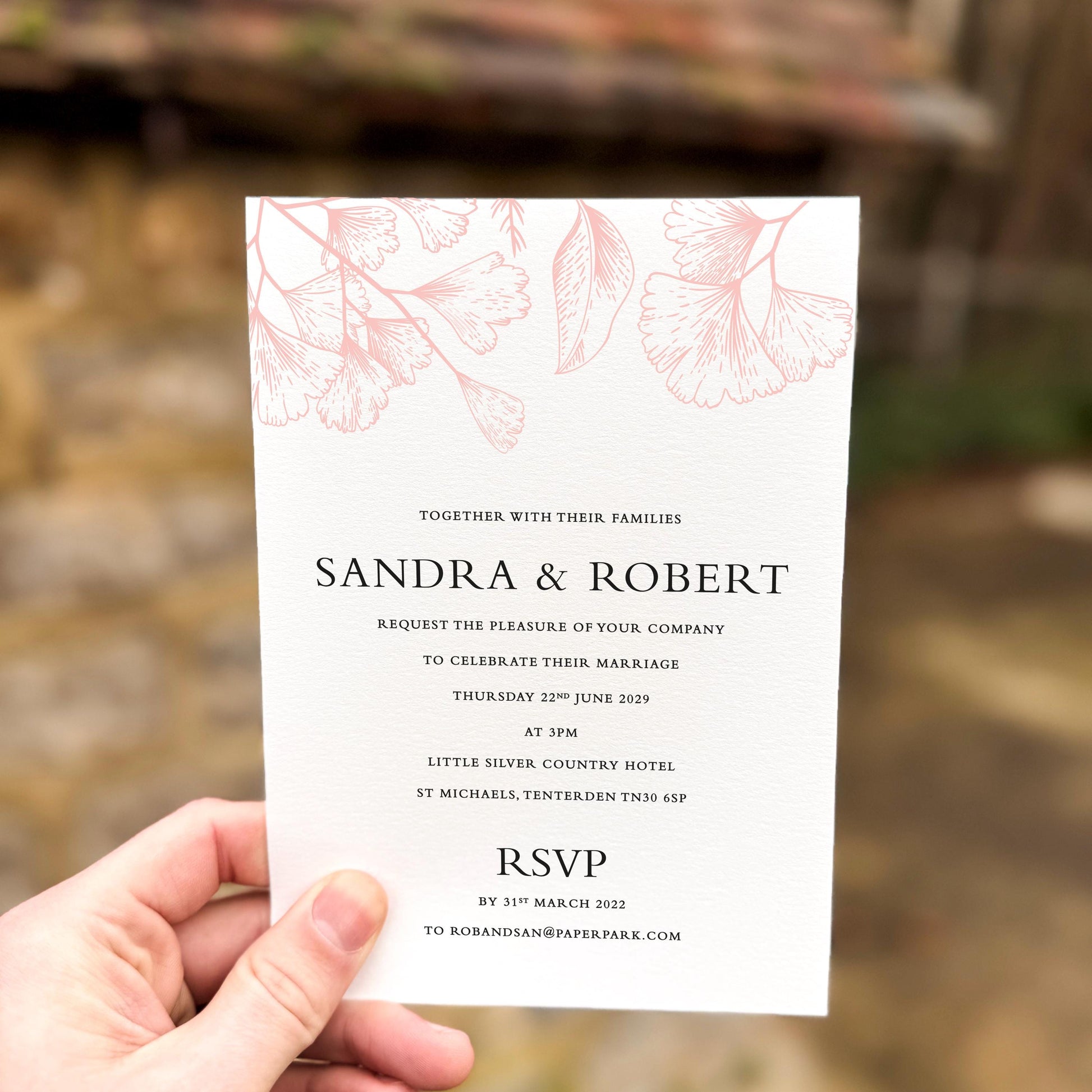 Hand holding a wedding invitation with soft peach line-art florals across the top, printed on textured white card.