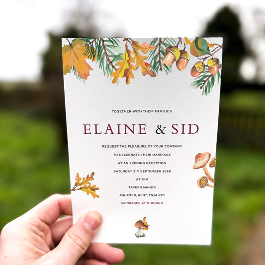 Hand holding an autumn wedding invitation decorated with illustrated oak leaves, acorns, pine branches and mushrooms around the edges.