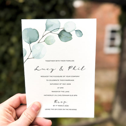 Hand holding a wedding invitation featuring soft green eucalyptus leaves painted in watercolour across the top of a textured card.
