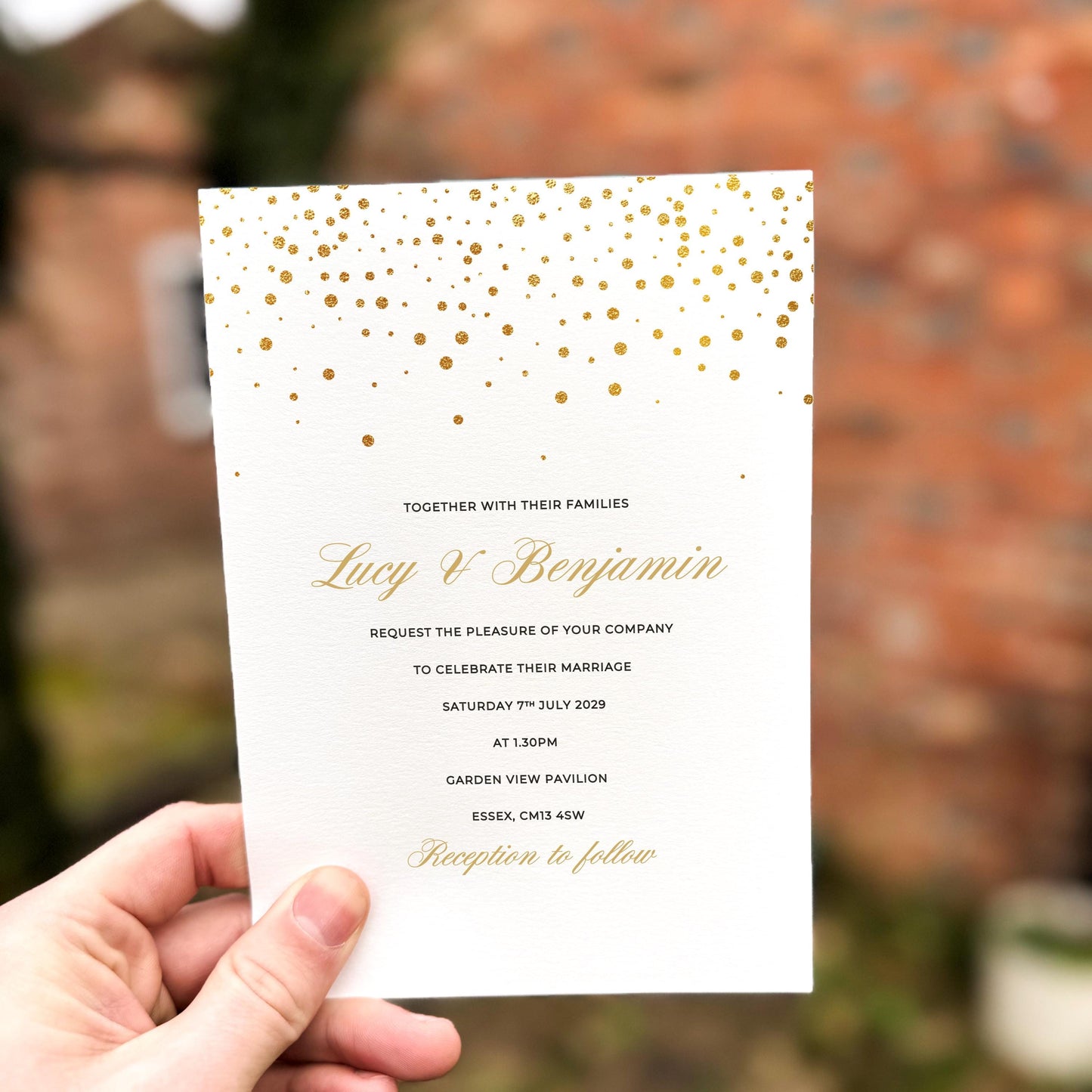 Hand holding a modern wedding invitation with scattered gold confetti dots across the top, printed on textured card.