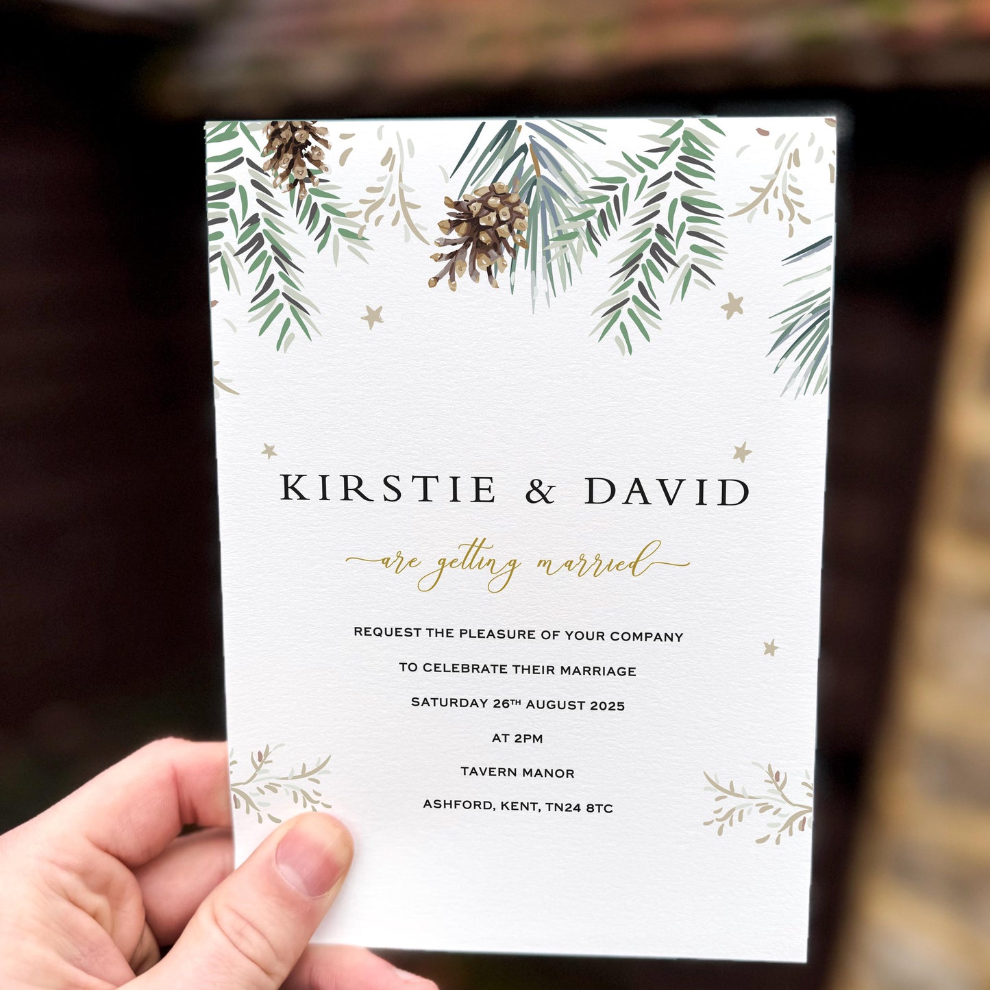 Hand holding a wedding invitation featuring painted pine branches, pinecones and subtle gold star accents on textured card.