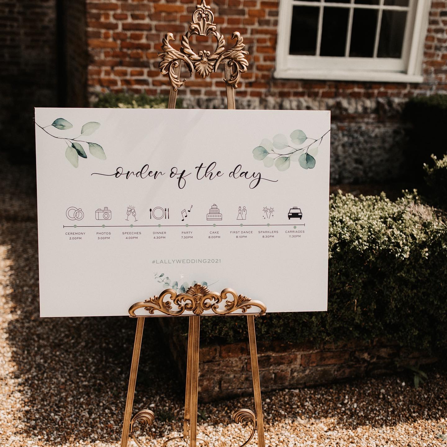 Elegant wedding ‘Order of the Day’ sign on a gold easel with eucalyptus illustrations, showing ceremony, photos, speeches, dinner, party, cake, first dance, sparklers and carriages timeline – outdoor venue setting